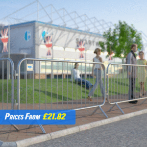 The image displays heavy-duty fixed leg barriers made of metal. These barriers have a series of vertical bars connected by two horizontal bars at the top and bottom, creating a sturdy grid structure. The legs are bent at right angles to provide stability on the ground. They are placed in a line, cordoning off an area outside where several people are gathered, suggesting an event or construction site in the background with flags and temporary structures visible.