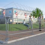 A series of interlocking metal pedestrian bar barriers create a queue outside an event with a group of people gathered behind them under clear skies during the day.