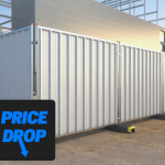 The image shows a series of vertical hoarding panels with a corrugated metal surface, set up around a construction site. The panels are supported by metal poles and concrete bases. In the foreground, there is a blue sign with bold white and yellow text that reads ‘PRICE DROP.