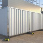 The image displays a series of galvanised metal hoarding panels with vertical stripes, set up in a straight line at a construction site. The panels are supported by yellow bases, and the background features scaffolding and a clear blue sky.