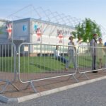 The image displays heavy-duty fixed leg barriers made of metal. These barriers have a series of vertical bars connected by two horizontal bars at the top and bottom, creating a sturdy grid structure. The legs are bent at right angles to provide stability on the ground. They are placed in a line, cordoning off an area outside where several people are gathered, suggesting an event or construction site in the background with flags and temporary structures visible.