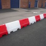 A series of alternating red and white plastic barriers, known as Evo barriers, set up in a straight line across a tarmac surface. The barriers are designed to demarcate a boundary or restrict access to the area behind them, which includes multiple industrial blue doors on buildings with brick walls. The image is taken during daylight with no visible people, emphasizing the barriers’ role in safety and organization in what appears to be an industrial or commercial setting.