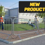 The image displays trade fixed leg barriers made of metal. These barriers have a series of 7 vertical bars connected by two horizontal bars at the top and bottom, creating a sturdy grid structure. The legs are bent at right angles to provide stability on the ground. They are placed in a line, cordoning off an area outside where several people are gathered, suggesting an event or construction site in the background with flags and temporary structures visible.