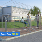 The image displays trade fixed leg barriers made of metal. These barriers have a series of 7 vertical bars connected by two horizontal bars at the top and bottom, creating a sturdy grid structure. The legs are bent at right angles to provide stability on the ground. They are placed in a line, cordoning off an area outside where several people are gathered, suggesting an event or construction site in the background with flags and temporary structures visible.