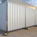 The image shows a series of vertical hoarding panels with a corrugated metal surface, set up around a construction site.